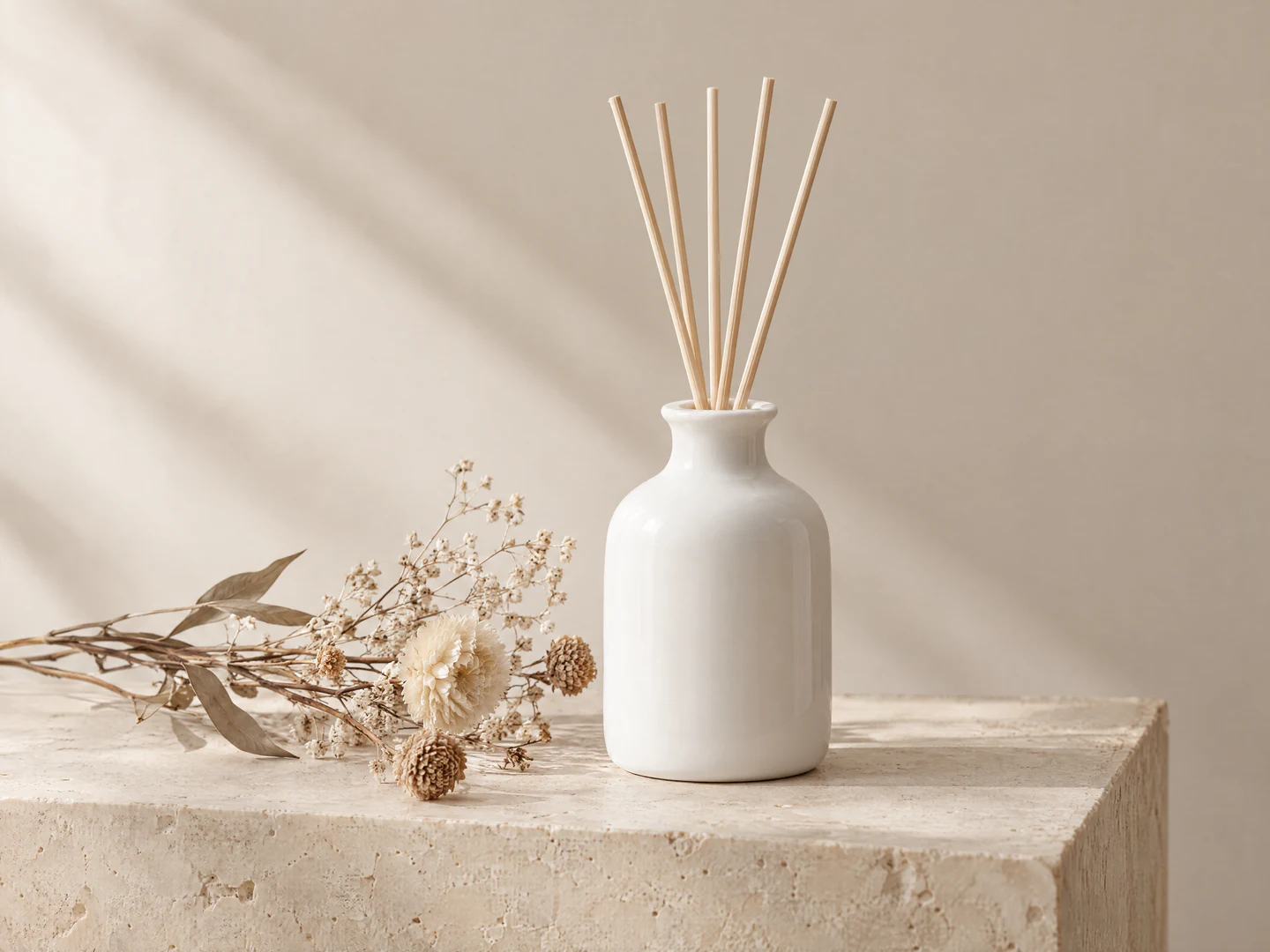 Foto de producto generada con IA: un difusor de varillas de cerámica blanca y flores secas sobre un bloque de piedra beige, con luz lateral suave que proyecta sombras en la pared